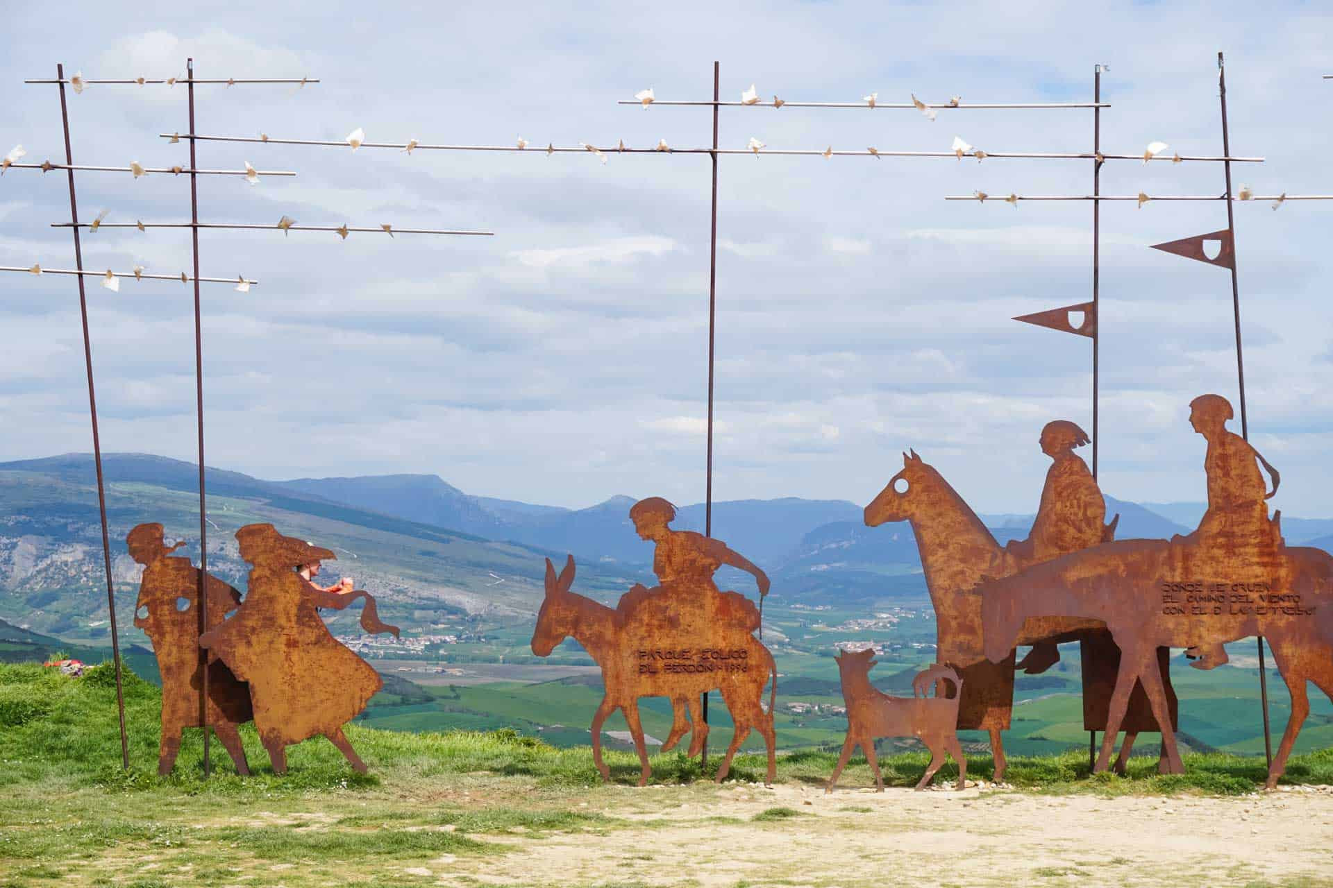 A group of seven Andaspain guides leap into the air with arms raised on a green mountain meadow, with rocky peaks, forested hillsides, and a dramatic cloudy sky behind them, and a cow grazing in the background.