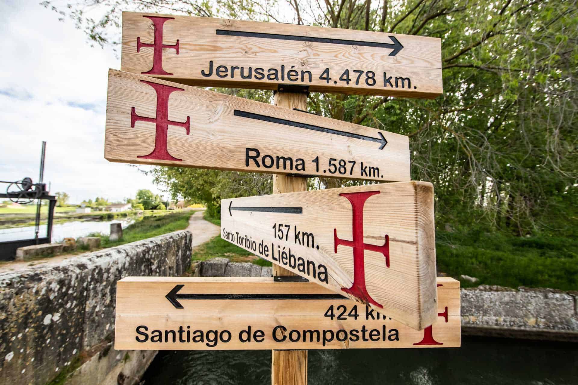Wooden signpost showing distances to Jerusalem, Rome, Santo Toribio de Liébana, and Santiago de Compostela with red cross symbols.