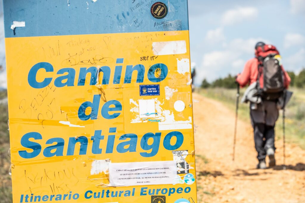 Yellow and blue Camino de Santiago sign beside a dirt path with a pilgrim walking away carrying a backpack.