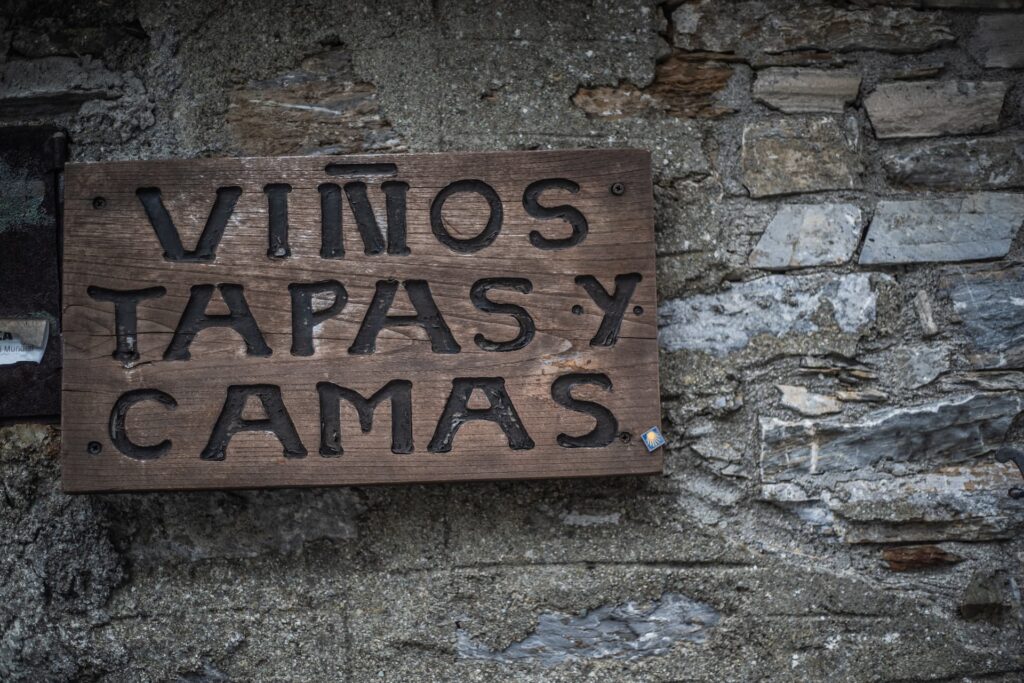 Wooden sign on a stone wall reading “Vinos Tapas y Camas,” offering wine, food, and lodging to pilgrims.