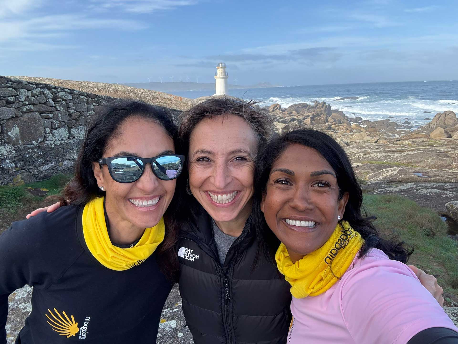 Three women smile warmly for a selfie wearing yellow Andaspain buffs, with a rocky coastline, lighthouse and crashing waves behind them.
