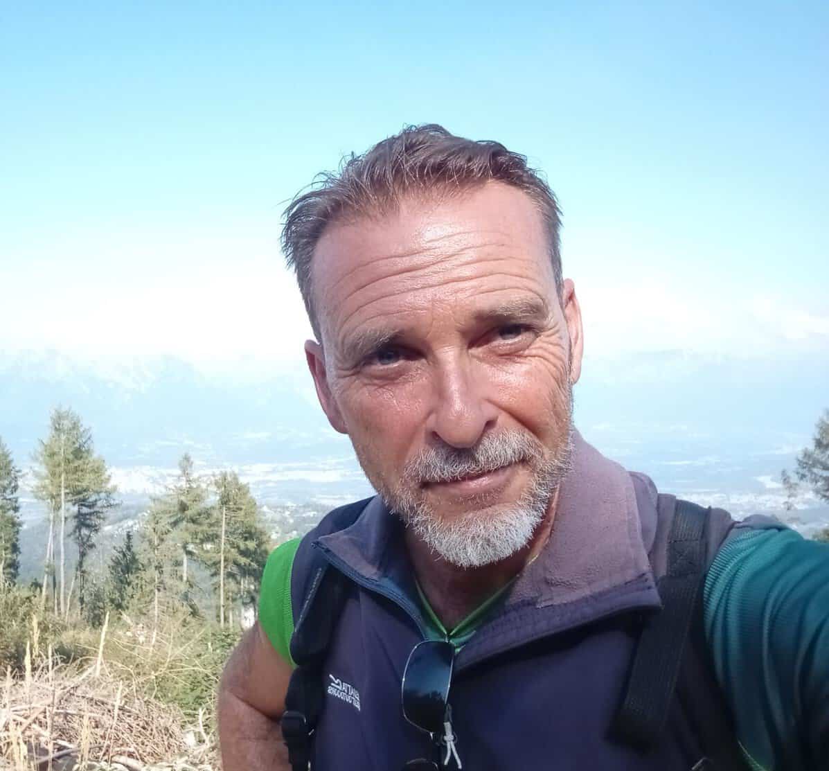 Emanuele Pierobon, Andaspain guide, takes a selfie on a hilltop wearing a gilet and backpack, with a hazy mountain valley stretching out behind him under a pale blue sky.