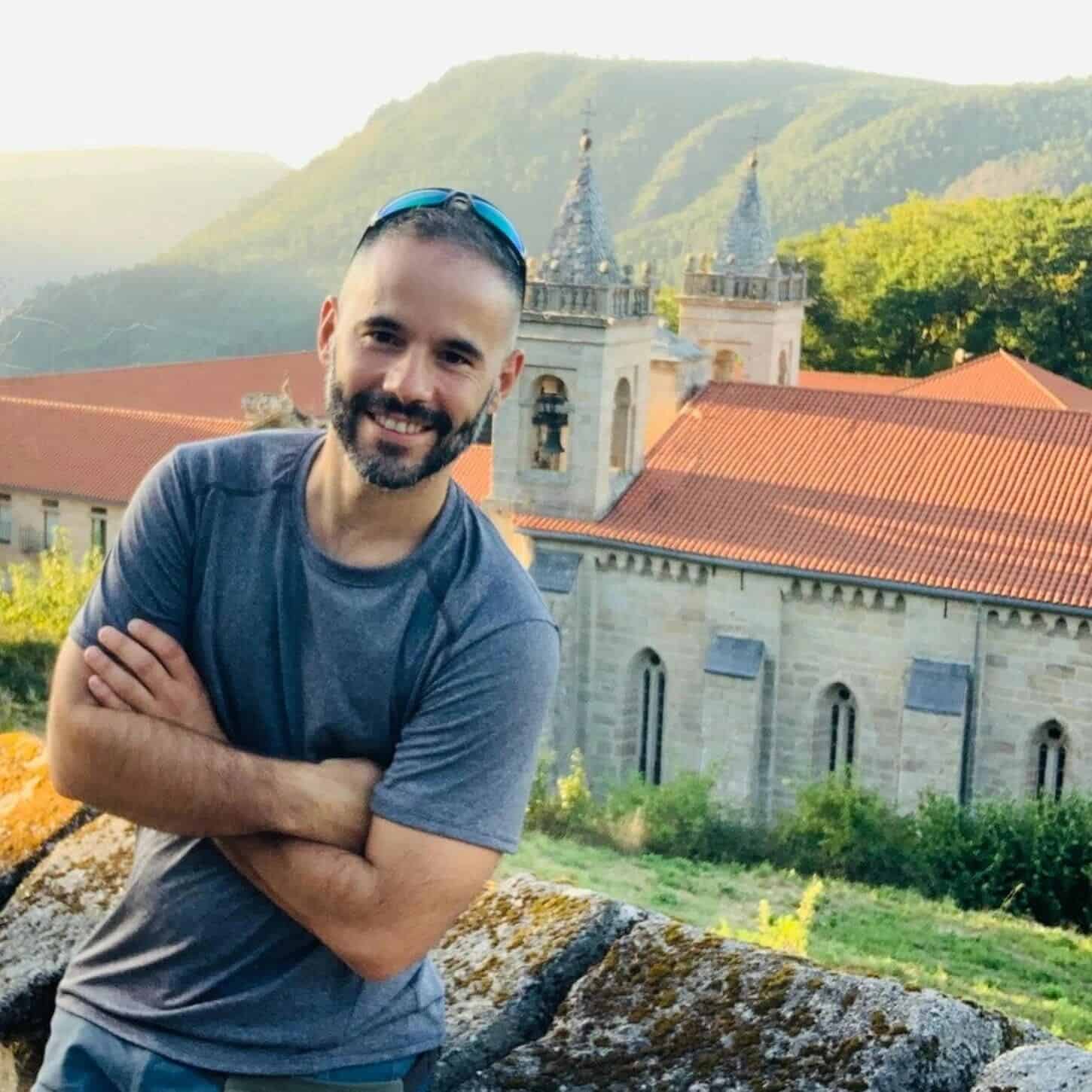 Miguel Calvo, Andaspain guide, smiles with arms crossed in front of a stone monastery with terracotta rooftops and green forested hills glowing in warm evening light behind him.