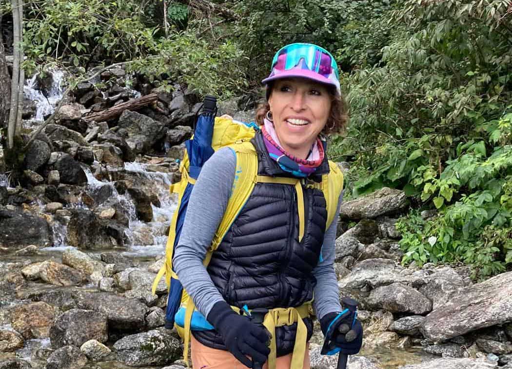 Azucena Collar, Andaspain guide, smiles confidently on a rocky mountain trail, fully equipped with a yellow and navy rucksack, hiking poles and a colourful cap, with a cascading waterfall and lush green woodland behind her.